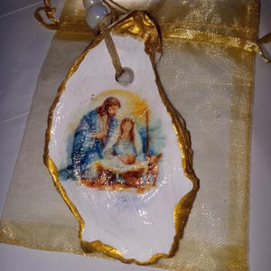 May include: A white oyster shell ornament with a gold trim, featuring a painted image of the nativity scene. The ornament is hanging from a gold string with white beads.