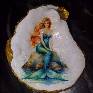 May include: A mermaid with long red hair sits on a rock with a blue and green tail. The mermaid is painted on a white oyster shell with a gold rim.