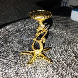 May include: A gold-colored metal candle holder featuring a base shaped like a five-pointed starfish. The central stem is formed by a vertical seahorse figure, which supports a small, circular, textured dish at the top for holding a candle.