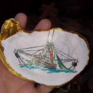 May include: A hand-painted oyster shell with a gold rim, featuring a detailed illustration of a fishing boat with nets, set against a blue water background.