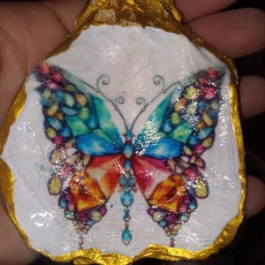 May include: A hand-painted oyster shell featuring a colorful butterfly design. The butterfly has blue, green, orange, and yellow wings. The shell is white inside with a gold-painted edge, creating a decorative piece.
