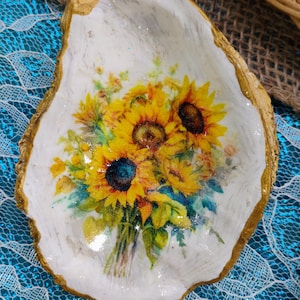 May include: A white oyster shell with a gold rim, decorated with a decoupage image of a bouquet of sunflowers. The shell is on a blue and white lace background.