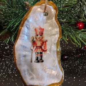 May include: A Christmas ornament made from a shell, featuring a painted image of a nutcracker soldier. The shell has a gold-painted border and a string for hanging. The ornament is set against a backdrop of evergreen branches and red berries.