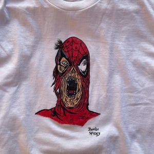 May include: White t-shirt featuring a red and black illustration of a zombie Spider-Man. The artwork depicts a decaying face with an open mouth. The text "Zombi Spidey" is written in black script below the image. The shirt is size M.
