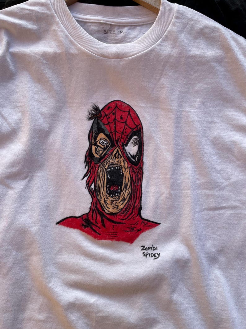 May include: White t-shirt featuring a red and black illustration of a zombie Spider-Man. The artwork depicts a decaying face with an open mouth. The text "Zombi Spidey" is written in black script below the image. The shirt is size M.