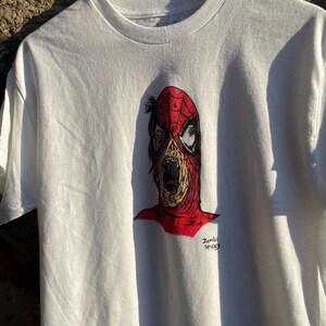May include: White t-shirt featuring a zombie Spider-Man graphic. The illustration depicts a decaying face with a red mask and open mouth. The text "Zombie Spidey" is printed below the image.