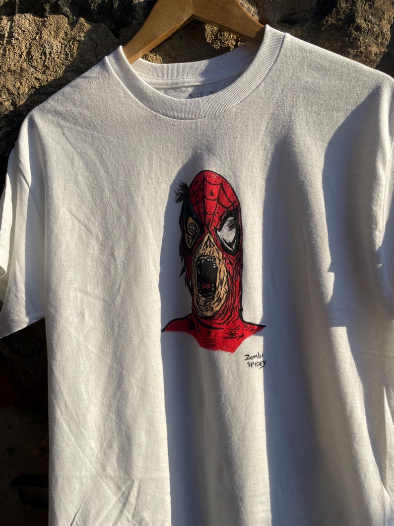 May include: White t-shirt featuring a zombie Spider-Man graphic. The illustration depicts a decaying face with a red mask and open mouth. The text "Zombie Spidey" is printed below the image.
