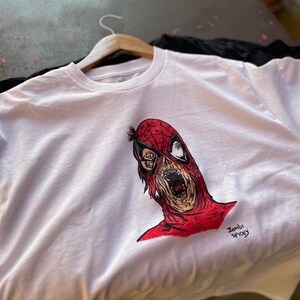 May include: White t-shirt featuring a red and black illustration of a zombie Spider-Man. The artwork includes the text "Zombi Spidey" in black. The shirt is hanging on a wooden hanger.