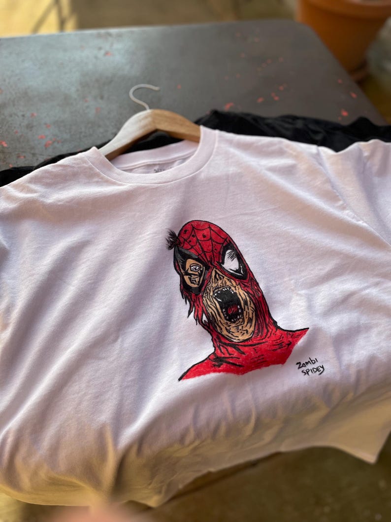 May include: White t-shirt featuring a red and black illustration of a zombie Spider-Man. The artwork includes the text "Zombi Spidey" in black. The shirt is hanging on a wooden hanger.