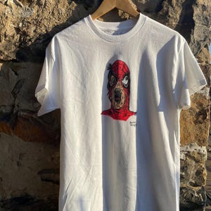 May include: White t-shirt featuring a graphic of a zombie-like character with a red and black mask. The design includes the text "Zombie Spidey". The shirt is hanging on a wooden hanger against a stone wall.