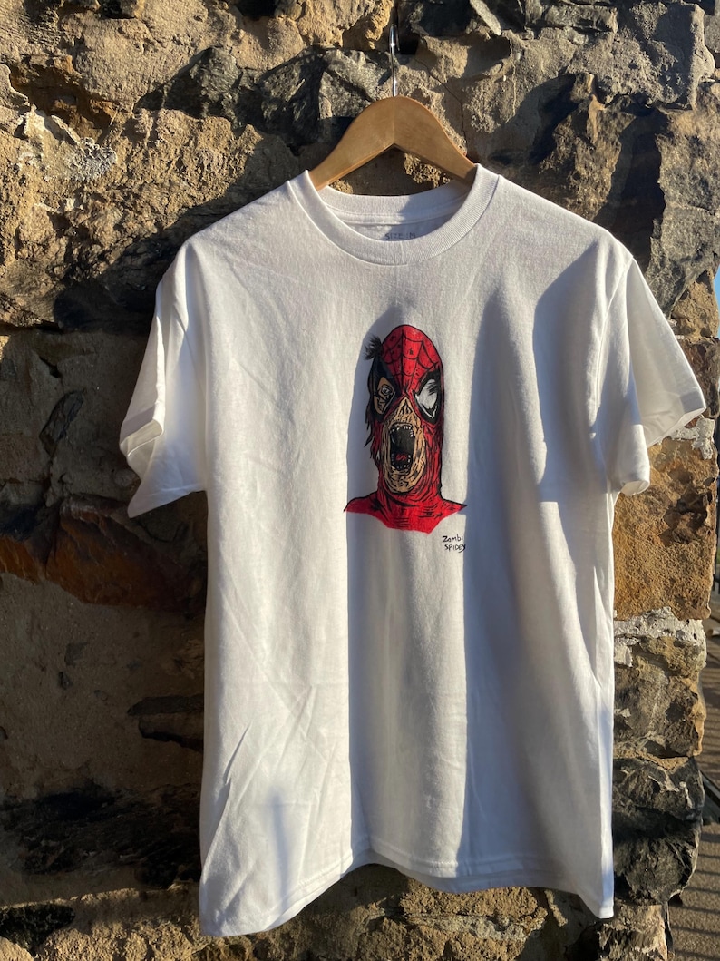 May include: White t-shirt featuring a graphic of a zombie-like character with a red and black mask. The design includes the text "Zombie Spidey". The shirt is hanging on a wooden hanger against a stone wall.