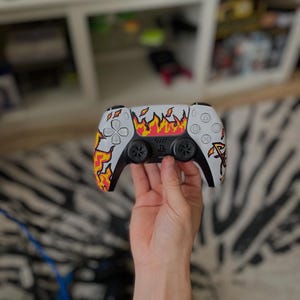 May include: A white and black PlayStation 5 controller featuring a flame design in yellow, orange, and red. The controller has black joysticks and buttons, with the PlayStation logo in pink.