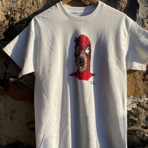 May include: White t-shirt featuring a graphic of a zombie-like Spider-Man. The design includes a red and black mask with exposed teeth and a red outline. The text "Zombie Spidey" is printed in black. The shirt is hanging on a wooden hanger.