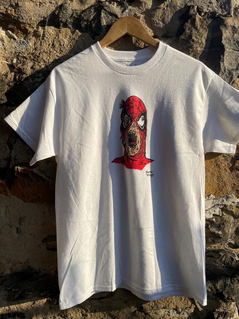 May include: White t-shirt featuring a graphic of a zombie-like Spider-Man. The design includes a red and black mask with exposed teeth and a red outline. The text "Zombie Spidey" is printed in black. The shirt is hanging on a wooden hanger.