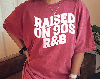 May include: A maroon t-shirt with the text "RAISED ON 90S R&B" in white, stylized lettering. The shirt has short sleeves and a relaxed fit. The image shows the shirt on a person.
