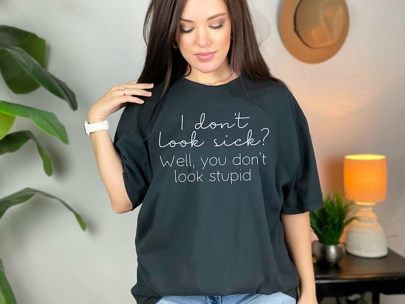 May include: Black t-shirt with the text "I don't look sick? Well, you don't look stupid" in a white, handwritten-style font. The shirt is a relaxed fit and has short sleeves.
