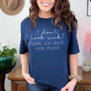 May include: Navy blue t-shirt with the text "I don't look sick? Well, you don't look stupid" in white script. The shirt is a crew neck style and appears to be made of a soft material.
