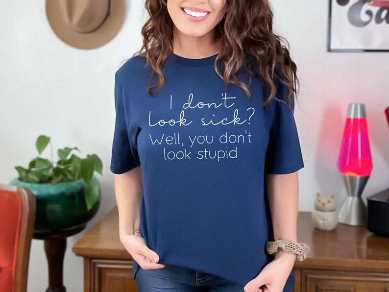 May include: Navy blue t-shirt with the text "I don't look sick? Well, you don't look stupid" in white script. The shirt is a crew neck style and appears to be made of a soft material.