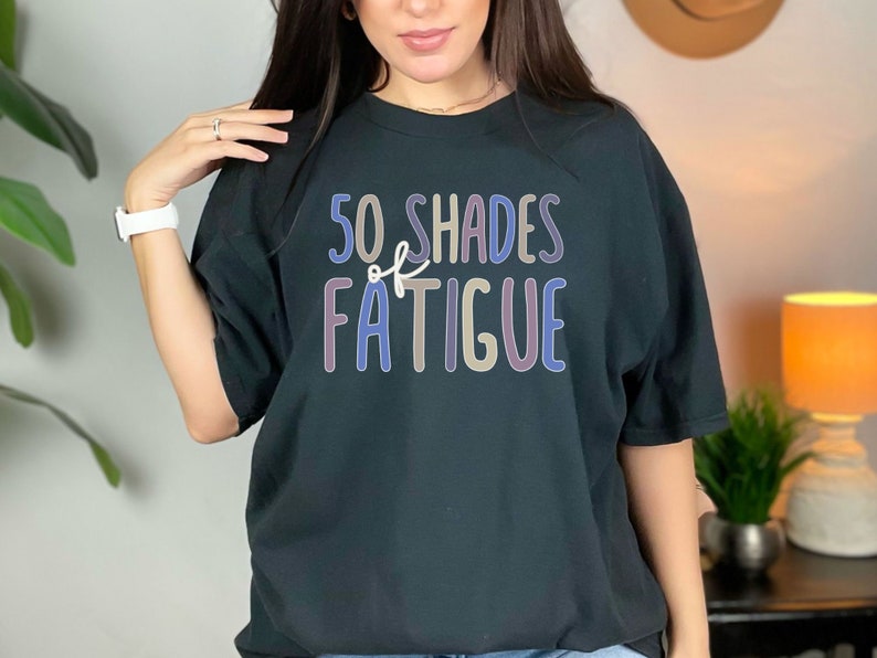 May include: Black t-shirt with the text "50 Shades of Fatigue" in a colorful, playful font. The shirt is a relaxed fit and appears to be made of a soft material. The design is centered on the front of the shirt.