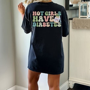 Może przedstawiać: Czarny, oversize'owy T-shirt z napisem "HOT GIRLS HAVE DIABETES" w kolorowej, retro czcionce. Obok tekstu znajduje się mały rysunek animowany butelki z insuliną.