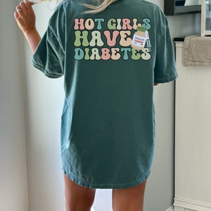 Może przedstawiać: Koszulka w kolorze turkusowym z napisem "HOT GIRLS HAVE DIABETES" w kolorowych literach. Obok tekstu znajduje się kresk&oacute;wkowa ilustracja butelki insuliny.