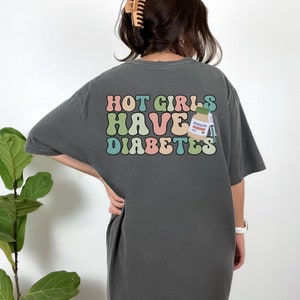 Może przedstawiać: Szary T-shirt z grafiką w stylu retro, na kt&oacute;rej widnieje napis "HOT GIRLS HAVE DIABETES" w kolorowych literach. Obok tekstu znajduje się kresk&oacute;wkowa ilustracja butelki insuliny i strzykawki.