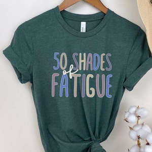 May include: A dark green t-shirt with the text "50 SHADES of FATIGUE" in colorful, outlined letters. The shirt is hanging on a wooden hanger. A straw hat and cotton stems are in the background.