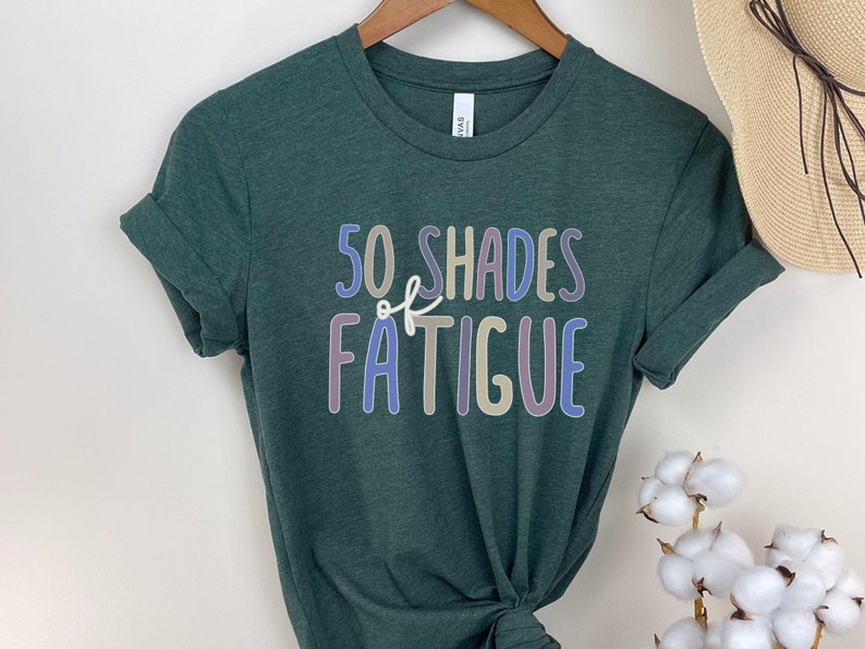 May include: A dark green t-shirt with the text "50 SHADES of FATIGUE" in colorful, outlined letters. The shirt is hanging on a wooden hanger. A straw hat and cotton stems are in the background.