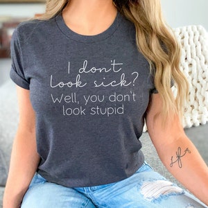 May include: Dark gray t-shirt with the text "I don't look sick? Well, you don't look stupid" in a white script font. The shirt has rolled-up sleeves and is made of a soft material. The shirt is a casual wear item.