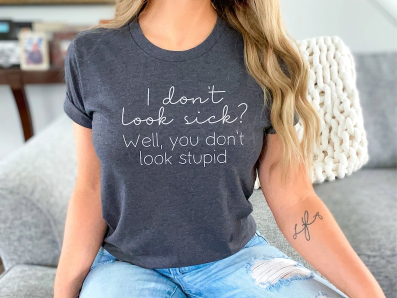 May include: Dark gray t-shirt with the text "I don't look sick? Well, you don't look stupid" in a white script font. The shirt has rolled-up sleeves and is made of a soft material. The shirt is a casual wear item.