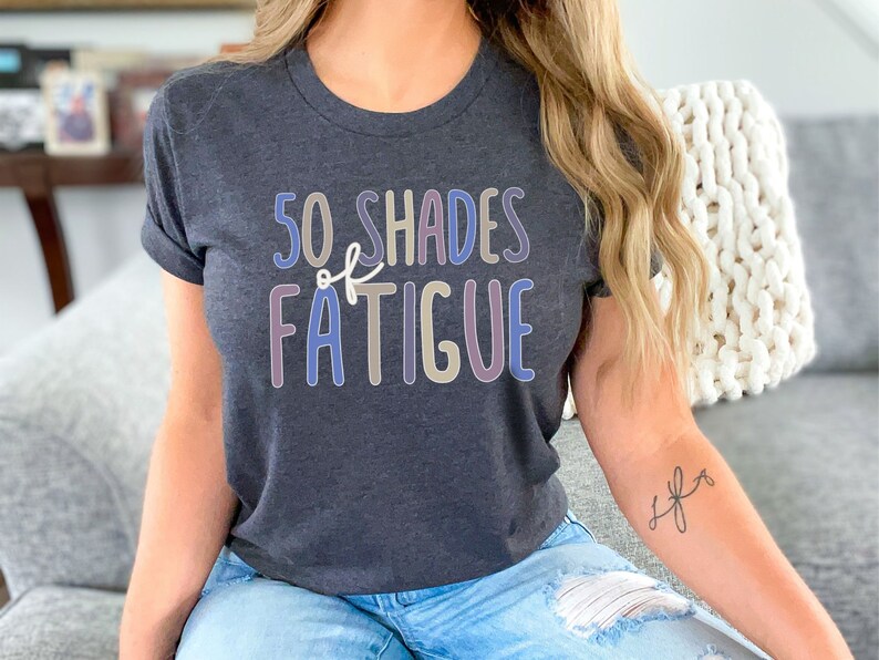 May include: Dark gray t-shirt with the text "50 Shades of Fatigue" in a colorful, playful font. The shirt has rolled-up sleeves and is made of a soft material. The overall theme is casual wear.