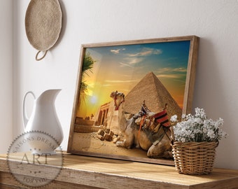 May include: A framed print depicting a camel resting in front of the Great Pyramid of Giza at sunset. The image features warm colors, with the pyramid and camel in the foreground and a vibrant sky in the background. The print is displayed on a wooden surface.