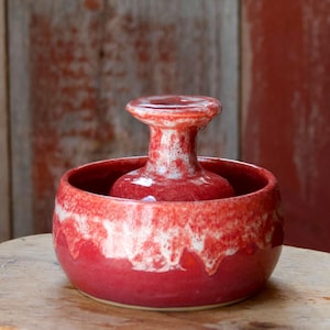 May include: A handmade, red ceramic bowl with a central, raised, vase-like element. The bowl and vase have a glossy finish with white accents, creating a unique, artistic look. The piece sits on a wooden surface, showcasing its handcrafted nature.