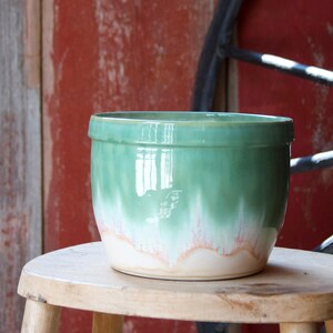 May include: A cylindrical ceramic bowl featuring a glossy glaze that transitions from a deep sage green at the top rim to a lighter mint green, ending in a creamy white base with subtle pinkish-orange drip patterns along the bottom edge.