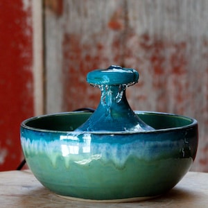 May include: A ceramic pet water fountain featuring a round bowl with a glossy glaze that transitions from a deep teal at the top rim to a lighter sage green at the base. A central spout allows water to flow down into the basin below.