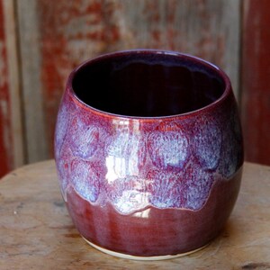 May include: A rounded ceramic mug featuring a glossy finish with a deep burgundy base color that transitions into a mottled, light lavender-blue glaze near the rim. The vessel has a wide, open mouth and a smooth, curved body with no handle attached.