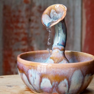May include: A ceramic water fountain with a unique design. The fountain features a bowl with a brown and purple glaze, and a slender, curved spout with water flowing from it. The spout and bowl have a glossy finish.