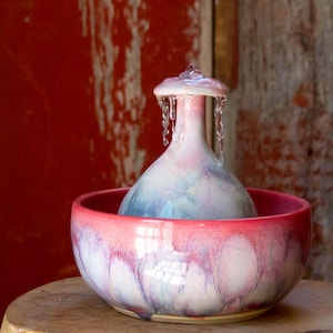 May include: A ceramic pet water fountain featuring a rounded basin and a central dome-shaped spout. The glaze displays a blend of soft pink, white, and light blue hues. Water flows from the top of the central spout, cascading down the sides.