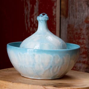 May include: A ceramic water fountain with a light blue and white glaze. The fountain features a bulbous top with a spout, set within a matching bowl. The design is simple and elegant, suitable for indoor decor.