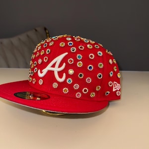 Puede incluir: Gorra de béisbol roja con visera plana, adornada con numerosos brillantes de colores. La letra blanca "A" está en la parte delantera, con un logotipo de New Era. Un accesorio de moda llamativo.
