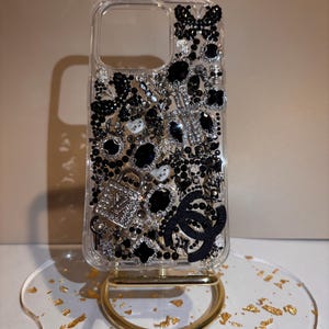 Puede incluir: Funda de teléfono transparente adornada con pedrería, cuentas y abalorios negros y plateados. El diseño incluye una cruz, un candado y un logotipo negro. La funda se exhibe sobre un soporte dorado con base transparente.