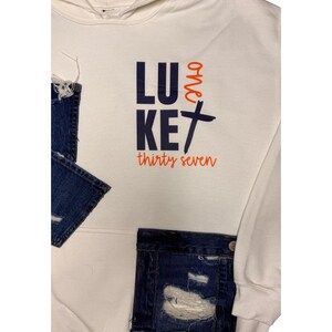 Puede incluir: Sudadera blanca con capucha y bolsillo de mezclilla. La parte delantera de la sudadera presenta las palabras "LUKE" y "one" en azul marino, con una cruz y las palabras "thirty seven" en naranja.