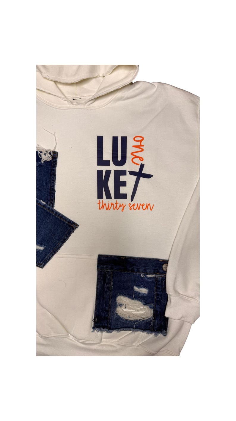 Puede incluir: Sudadera blanca con capucha y bolsillo de mezclilla. La parte delantera de la sudadera presenta las palabras "LUKE" y "one" en azul marino, con una cruz y las palabras "thirty seven" en naranja.