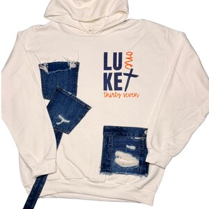 Puede incluir: Sudadera con capucha color crema con parches y detalles de mezclilla desgastada. La sudadera presenta el texto "LUKE thirty seven" en azul marino y naranja. El dise&ntilde;o incluye una capucha y un bolsillo de canguro.
