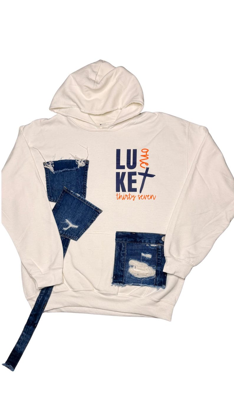 Puede incluir: Sudadera con capucha color crema con parches y detalles de mezclilla desgastada. La sudadera presenta el texto "LUKE thirty seven" en azul marino y naranja. El dise&ntilde;o incluye una capucha y un bolsillo de canguro.