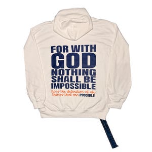 Puede incluir: Sudadera con capucha color crema con el texto "FOR WITH GOD NOTHING SHALL BE IMPOSSIBLE" en azul marino. La frase "He is the definition of all things that are POSSIBLE" est&aacute; impresa debajo en naranja. La sudadera tiene un cord&oacute;n y un bolsillo de canguro.