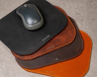 Könnte beinhalten: Ein Stapel personalisierter Leder-Mauspads in Schwarz, Braun und Orange. Eine graue kabellose Maus liegt auf dem obersten schwarzen Pad, das mit dem Namen "DAVID" graviert ist. Andere Pads sind mit Initialen graviert.
