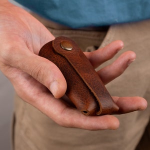 Könnte beinhalten: Ein brauner Leder-Schlüsselanhänger mit einem Metallknopfverschluss.