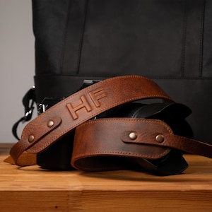 May include: A brown leather camera strap featuring the embossed letters HF, secured with metallic rivets. The strap is draped over a black camera body, with a black camera lens visible to the left and a black bag positioned in the background.