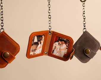 May include: Three leather keychain photo holders hanging from a wooden rod. The center holder is open, displaying two rectangular photographs of people. Each holder features a metal chain, a snap button closure, and visible stitching along the edges.